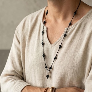 Op de afbeelding: Een lange zwarte kralenketting met een zilveren hanger. De ketting heeft meerdere zwarte kralen geregen aan een zwart koord. De hanger is een kleine zilveren driehoek. De persoon draagt een beige V-hals shirt.