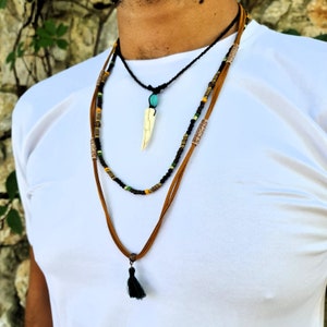 May include: Layered necklace with a brown leather cord, black cord, and beaded cord. The necklace features a bone pendant and a black tassel.
