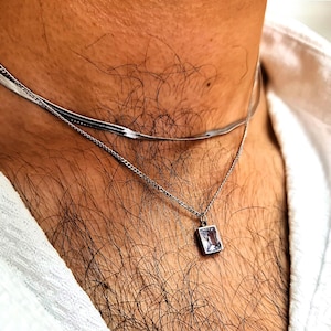 Könnte beinhalten: Eine silberne Kettenkette mit einem quadratischen Anhänger mit einem klaren Kristall. Die Kette ist über eine dünnere silberne Kette gelegt.