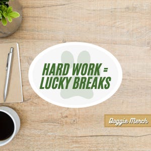 May include: White oval sticker with a green paw print and the text "Hard Work = Lucky Breaks". The sticker is on a wood surface. The text "Doggie Merch" is in a brown rectangle at the bottom of the image.