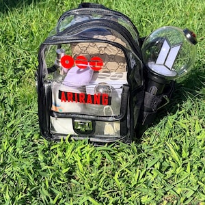 May include: A clear, black-trimmed backpack filled with various items, including a small purse, cosmetics, and a purple item. The backpack has the word "ARIRANG" in red lettering. A light stick with a white logo is attached to the side. The backpack is sitting on green grass.