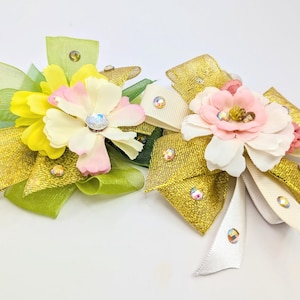 May include: Two handmade hair bows with a gold and green ribbon, white ribbon, and pink and yellow flowers. The bows are decorated with iridescent rhinestones.