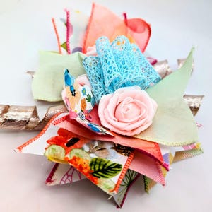 May include: A colorful handmade hair bow featuring a pink rose, blue lace, and various fabric scraps in shades of pink, orange, green, and floral patterns. A gold ribbon adds a touch of sparkle to this unique accessory.