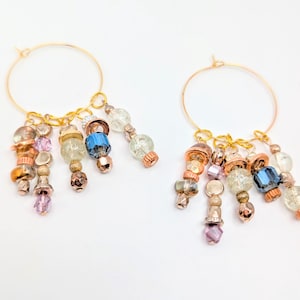 May include: Two gold hoop earrings with dangling beaded charms. The charms feature a mix of colors, including copper, blue, clear, and pink beads. The earrings are displayed on a white background.