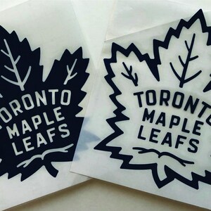 K&ouml;nnte beinhalten: Zwei Aufkleber mit dem Logo der Toronto Maple Leafs. Jeder Aufkleber zeigt ein stilisiertes Ahornblatt in Schwarz mit den Worten "TORONTO MAPLE LEAFS" in Wei&szlig;. Die Aufkleber befinden sich auf einer transparenten R&uuml;ckseite.