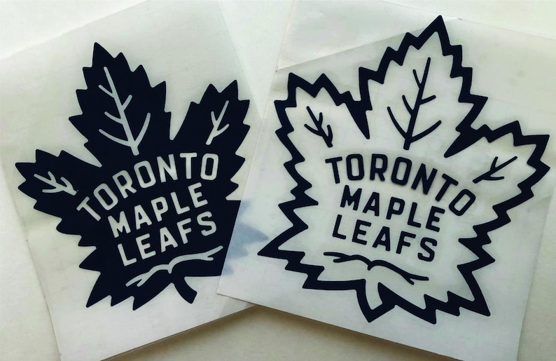 K&ouml;nnte beinhalten: Zwei Aufkleber mit dem Logo der Toronto Maple Leafs. Jeder Aufkleber zeigt ein stilisiertes Ahornblatt in Schwarz mit den Worten "TORONTO MAPLE LEAFS" in Wei&szlig;. Die Aufkleber befinden sich auf einer transparenten R&uuml;ckseite.