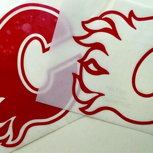K&ouml;nnte beinhalten: Zwei rot-wei&szlig;e Vinyl-Aufkleber mit dem Logo der Calgary Flames. Das Logo ist ein stilisiertes 'C' mit einem Flammendesign. Ein Aufkleber ist vollst&auml;ndig sichtbar, der andere ist teilweise bedeckt.