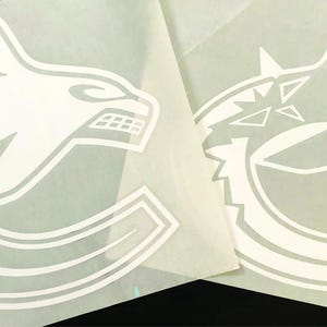 K&ouml;nnte beinhalten: Zwei wei&szlig;e Vinylaufkleber mit dem Logo der Vancouver Canucks. Das Logo ist ein stilisierter Orca-Walkopf, in dessen Design der Teamname integriert ist. Die Aufkleber befinden sich auf einer wei&szlig;en R&uuml;ckseite.