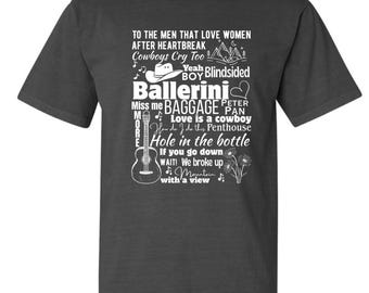 May include: Dark gray t-shirt with white text and graphics. The text includes song titles and lyrics, along with a guitar, cowboy hat, and floral elements. The shirt's design is centered on the front.