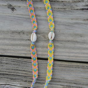 May include: Two friendship bracelets with a yellow, pink, blue and green color pattern. Each bracelet has a white seashell charm.