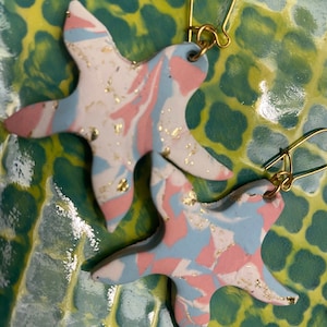 May include: A pair of pink, blue, and white marbled starfish earrings with gold flecks. The earrings are on a green and yellow patterned surface.