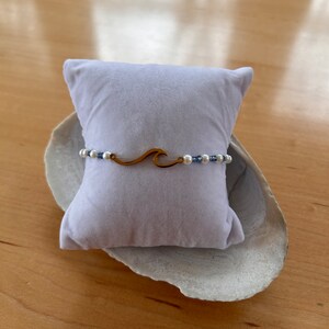 May include: A gold wave charm bracelet with white and blue beads on a white velvet cushion.