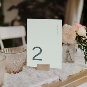 May include: A wedding table setting with a table number card. The card is light green with the number 2 in dark teal. The card is held by a small wooden block. A vase of flowers and two decorative glasses are also on the table.