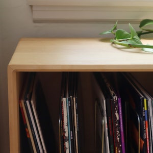 May include: A light wood record storage cabinet with a rounded top edge. The cabinet is filled with vinyl records with colorful album covers.