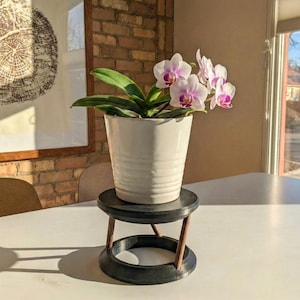 May include: A white ceramic pot with an orchid plant, featuring pink and purple flowers, sits on a small, black plant stand with brown supports. The stand is on a white table, with a brick wall and window in the background.