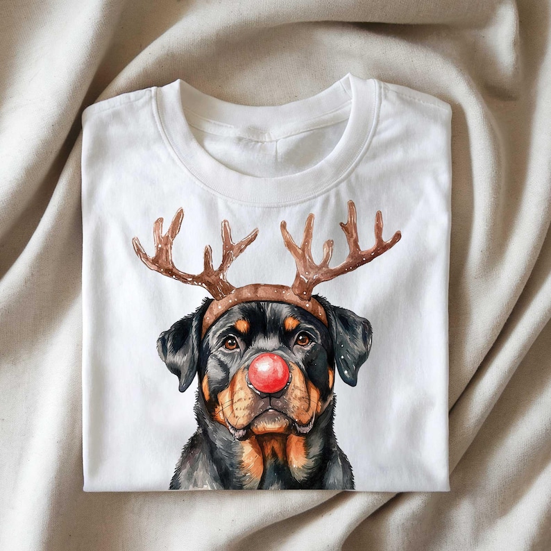 May include: White t-shirt featuring a Rottweiler dog wearing reindeer antlers and a red nose. The dog's fur is black and brown, with detailed watercolor-style artwork. The shirt is folded neatly on a neutral-colored surface.