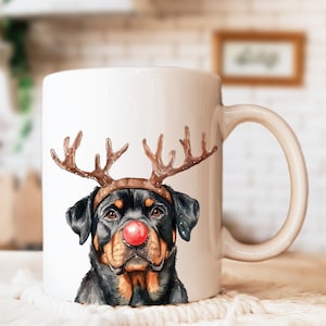 May include: White ceramic mug featuring a Rottweiler dog with a red nose and reindeer antlers. The dog's face is a watercolor illustration, with black, brown, and tan fur. The mug has a curved handle and is set against a blurred background.