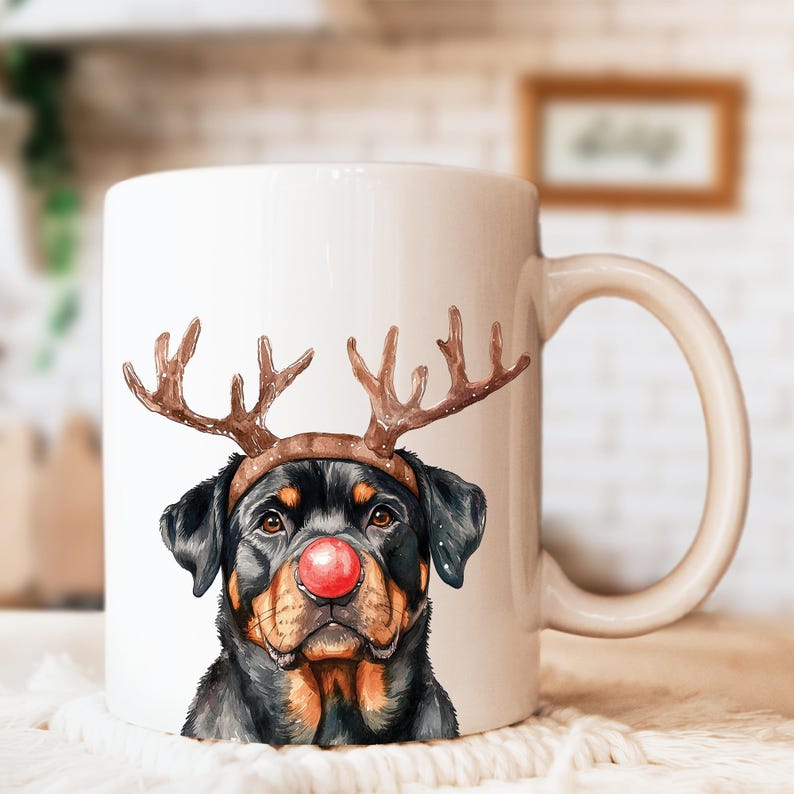 May include: White ceramic mug featuring a Rottweiler dog with a red nose and reindeer antlers. The dog's face is a watercolor illustration, with black, brown, and tan fur. The mug has a curved handle and is set against a blurred background.