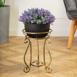 May include: A gold metal plant stand with a round top and three legs. A black planter with purple flowers sits on top of the stand.