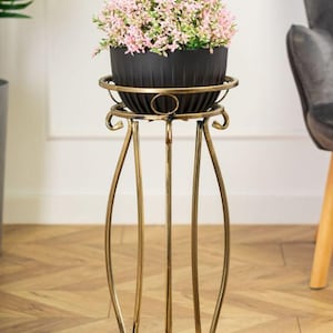 Könnte beinhalten: Ein goldfarbener Pflanzenständer aus Metall mit einer runden Oberseite und drei Beinen. Ein schwarzer Topf mit rosa Blumen steht auf dem Ständer.