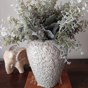 May include: A grey textured ceramic vase with a bouquet of light green eucalyptus branches. The vase is sitting on a brown book titled "The Mill on the Floss" by George Eliot.