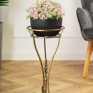 May include: A black plant pot with pink and green flowers on a decorative gold-coloured plant stand. The stand has a scrolled base and a twisted central support, adding elegance to the floral display. The stand is approximately 70 cm tall.