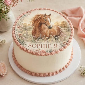 Könnte beinhalten: Ein runder Kuchen mit glatter weißer Glasur und rosa verzierten Rändern. Oben befindet sich ein essbares Bild eines braunen Pferdes auf einer Blumenwiese mit einem Holzzaun im Hintergrund. Der Text "SOPHIE 9" ist in braunen Buchstaben gedruckt.