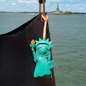 May include: A black handbag with a dark bamboo handle and a light peach leather strap attached to a teal leather charm shaped like the Statue of Liberty. The charm features a yellow flame and a book with the text JULY IV clearly visible.