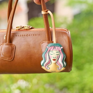 Puede incluir: Un bolso de mano de cuero marr&oacute;n con una cremallera dorada y un charm de cuero sujeto al asa. El charm es una ilustraci&oacute;n verde, rosa y blanca de una mujer con la palabra "VIRGO" escrita debajo.