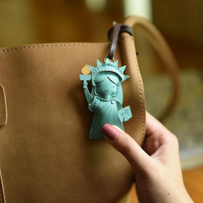May include: A teal Statue of Liberty charm with a brown leather strap. The charm is holding a torch and a tablet that reads "JULY IV". The charm is attached to a brown purse.
