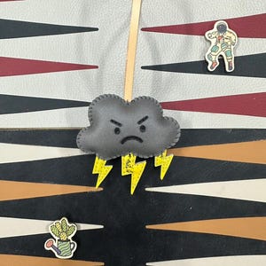 May include: A gray felt cloud with a frowning face and yellow lightning bolts hanging from a tan leather cord. The cloud is hanging against a black and white checkerboard background.
