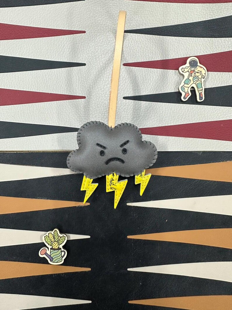 May include: A gray felt cloud with a frowning face and yellow lightning bolts hanging from a tan leather cord. The cloud is hanging against a black and white checkerboard background.