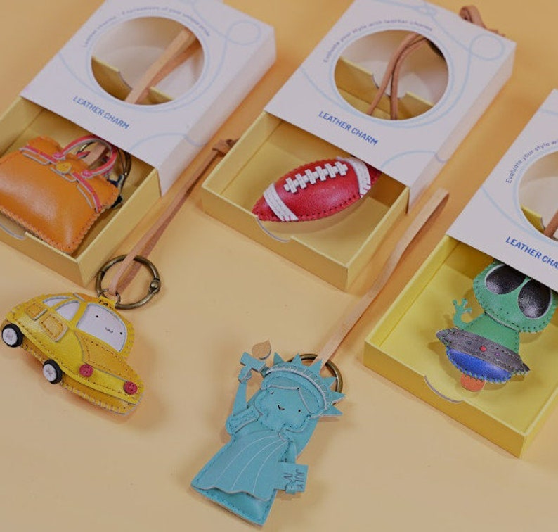 May include: A collection of leather charm keychains in gift boxes. The keychains include a yellow car, a red football, a blue Statue of Liberty, and a green alien. The boxes are white with the words "LEATHER CHARM".