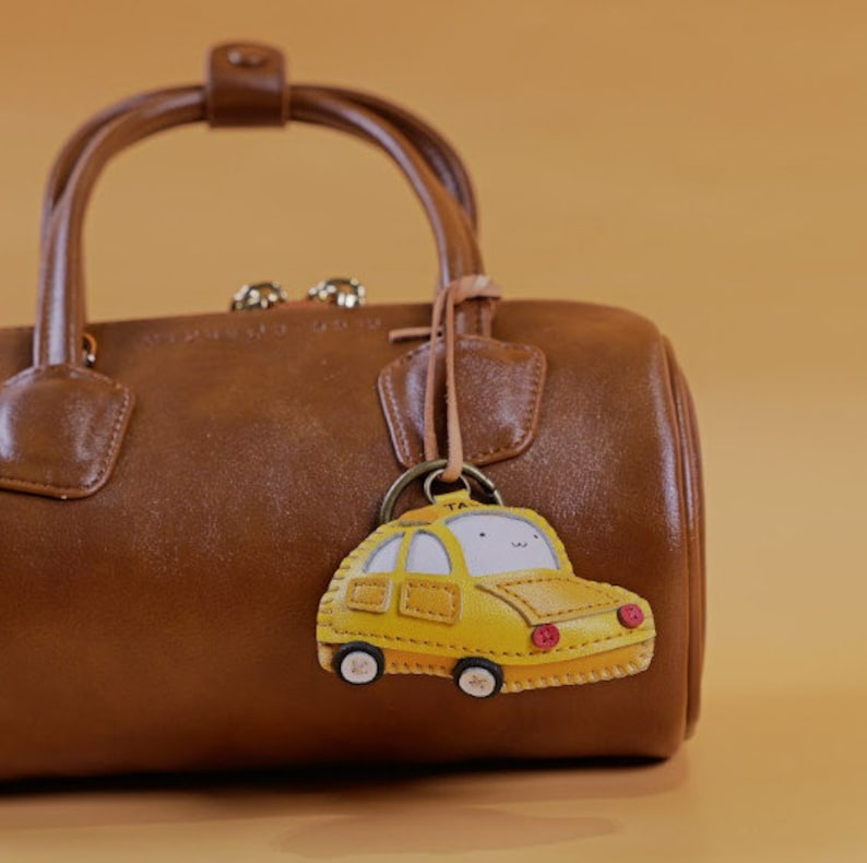 May include: A brown leather handbag with a top handle and a yellow taxi cab keychain. The keychain has a smiling face and the word "TAXI" on top. The bag is sitting on a light brown surface.