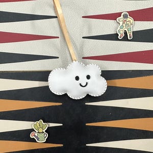 May include: A white felt cloud with a stitched smile hanging from a brown leather cord. The cloud is on a black and white patterned surface with a red and white patterned surface above it. There are two small wooden pins on the surface, one of an astronaut and one of a cactus in a pot.