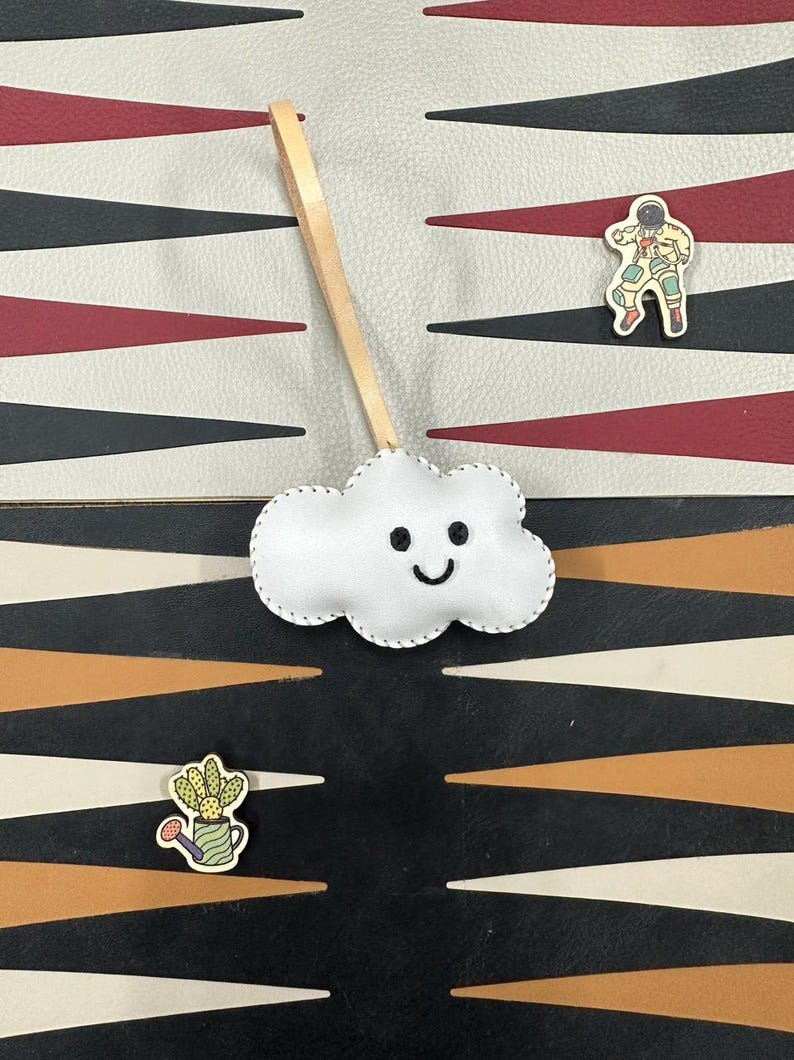May include: A white felt cloud with a stitched smile hanging from a brown leather cord. The cloud is on a black and white patterned surface with a red and white patterned surface above it. There are two small wooden pins on the surface, one of an astronaut and one of a cactus in a pot.