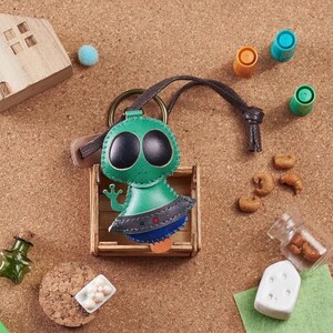 May include: A green and black leather alien keychain with large black eyes and a small wooden crate. The alien has a blue and gray skirt and a brown leather strap. The background includes small decorative items.