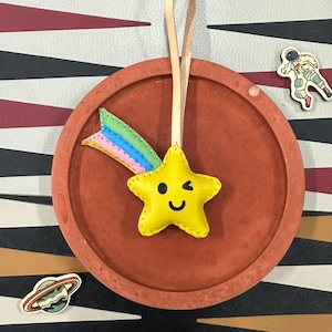 May include: A yellow star-shaped leather ornament with a smiling face and a winking eye. The star has a rainbow-colored leather tail attached to it. The ornament is hanging from a brown leather strap.