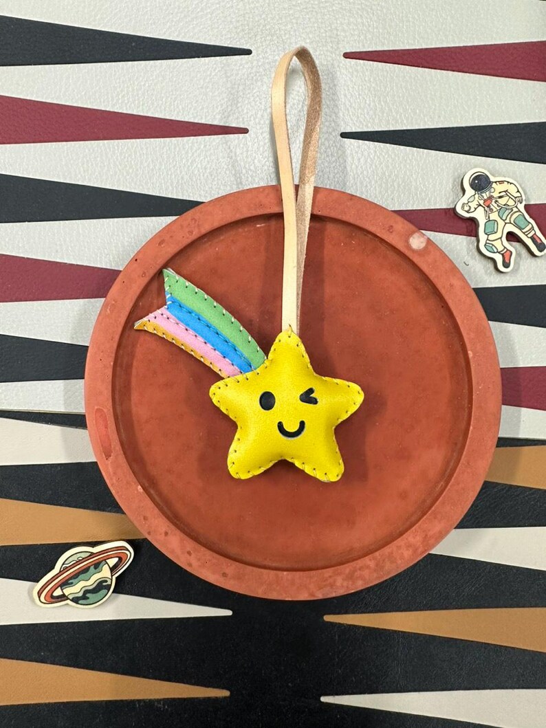 May include: A yellow star-shaped leather ornament with a smiling face and a winking eye. The star has a rainbow-colored leather tail attached to it. The ornament is hanging from a brown leather strap.