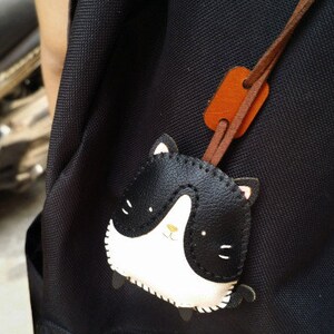 May include: A black backpack with a black and white leather cat charm attached to a brown leather strap.