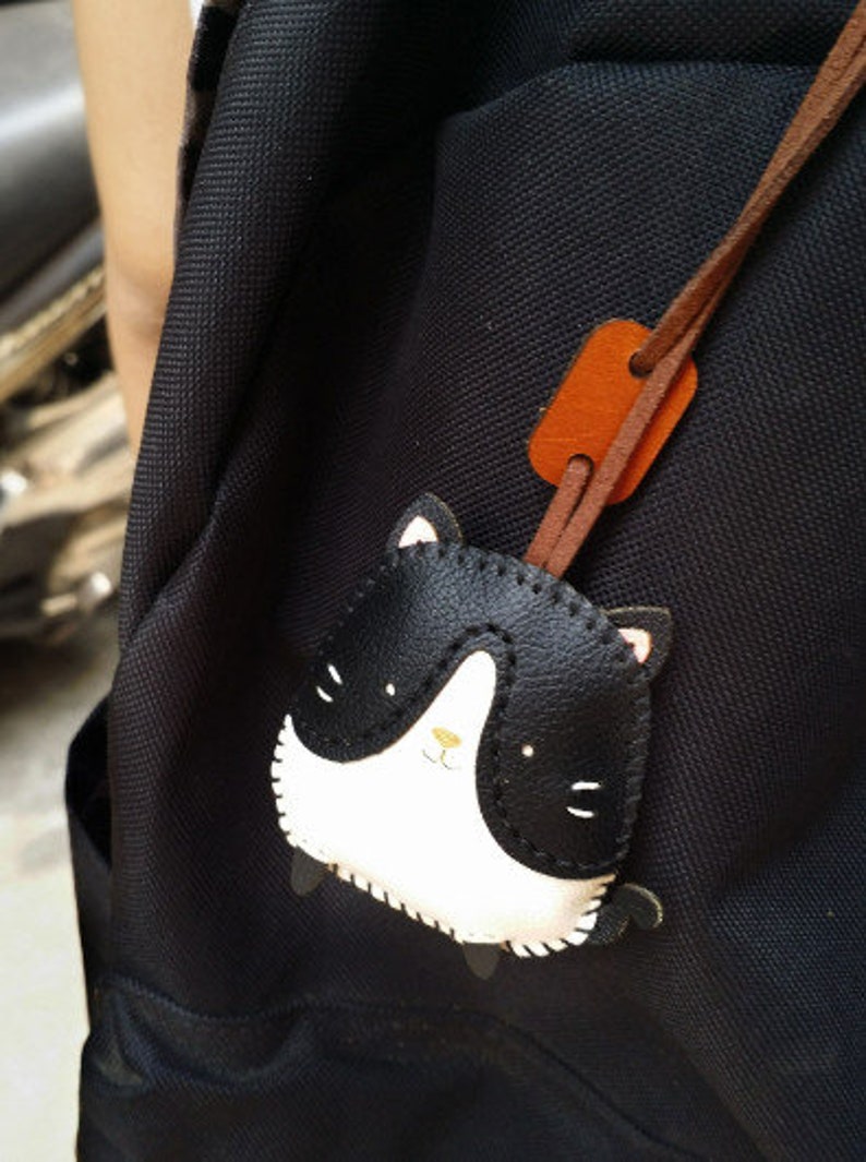 May include: A black backpack with a black and white leather cat charm attached to a brown leather strap.