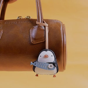 May include: A brown leather handbag with a penguin-shaped keychain. The keychain is black, white, and blue, holding a blue fish with a white X. The penguin has orange feet and a small orange beak.
