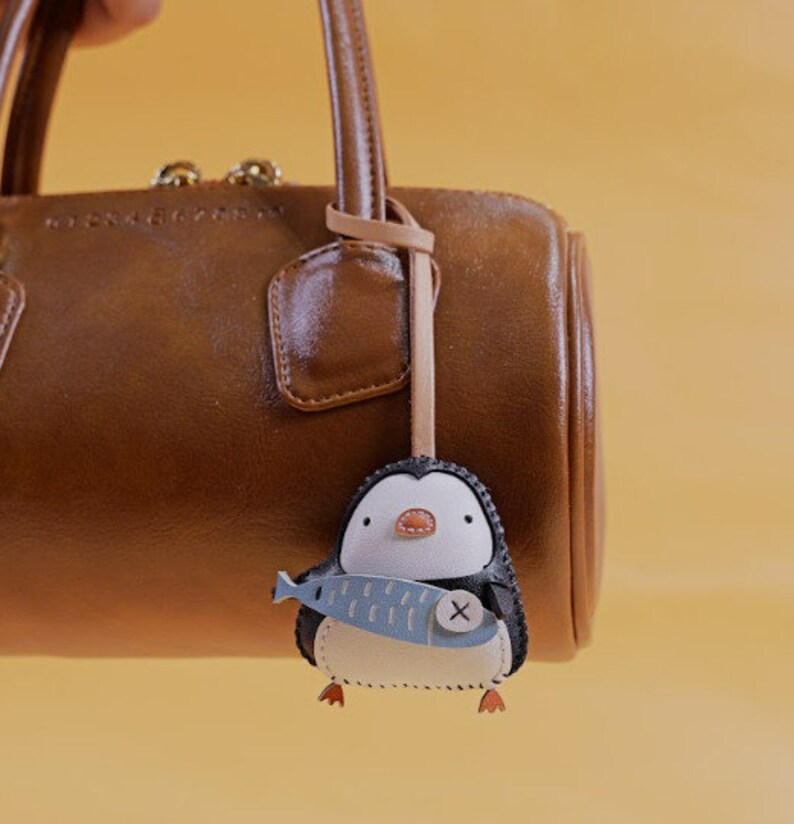 May include: A brown leather handbag with a penguin-shaped keychain. The keychain is black, white, and blue, holding a blue fish with a white X. The penguin has orange feet and a small orange beak.