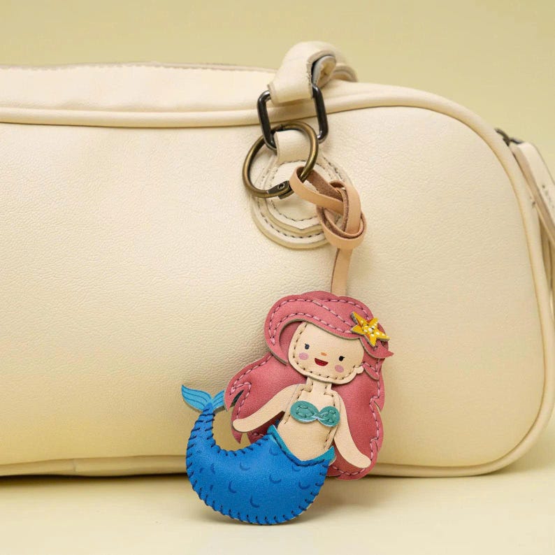 May include: A cream-colored handbag featuring a leather keychain charm shaped like a mermaid. The mermaid has long pink hair, a yellow starfish accessory, a teal seashell top, and a blue tail with scale details, attached by a metal ring.