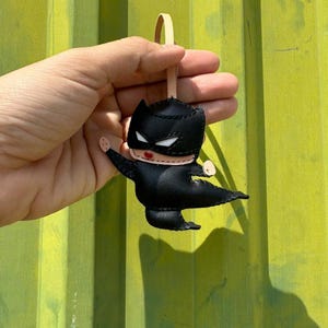 May include: A black superhero-themed keychain with a leather strap. The keychain features a stylized character with a black mask, cape, and body. The character has white eyes and red lips. The background is a textured, olive-green surface.