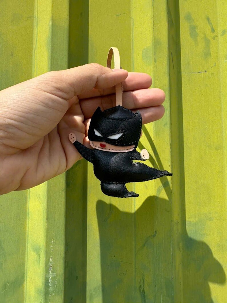 May include: A black superhero-themed keychain with a leather strap. The keychain features a stylized character with a black mask, cape, and body. The character has white eyes and red lips. The background is a textured, olive-green surface.