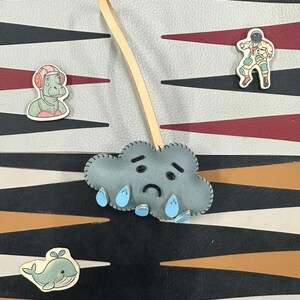 May include: A gray felt cloud with a sad face and blue raindrops hanging from it. The cloud is attached to a brown leather strap. The cloud is on a black and white patterned background.