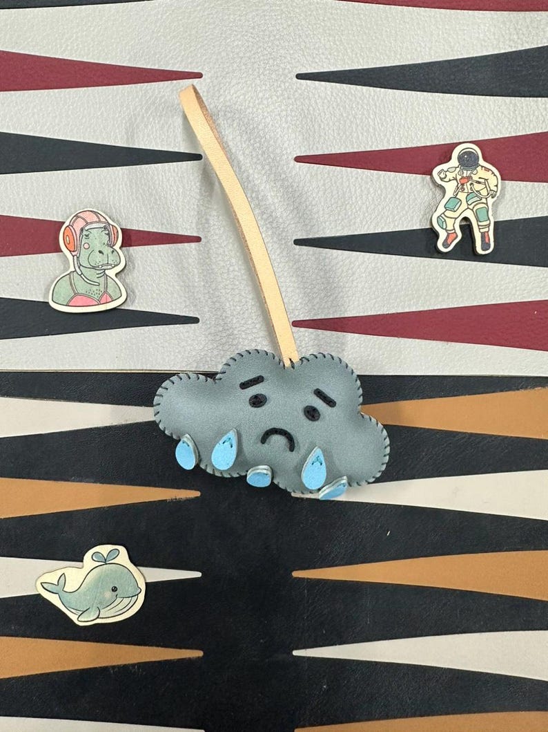 May include: A gray felt cloud with a sad face and blue raindrops hanging from it. The cloud is attached to a brown leather strap. The cloud is on a black and white patterned background.