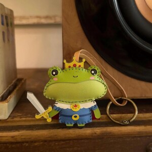May include: A handmade felt frog ornament, wearing a blue and gold outfit with a red cape and a gold crown. The frog holds a silver sword. The ornament has a leather loop for hanging and is set against a wooden background.