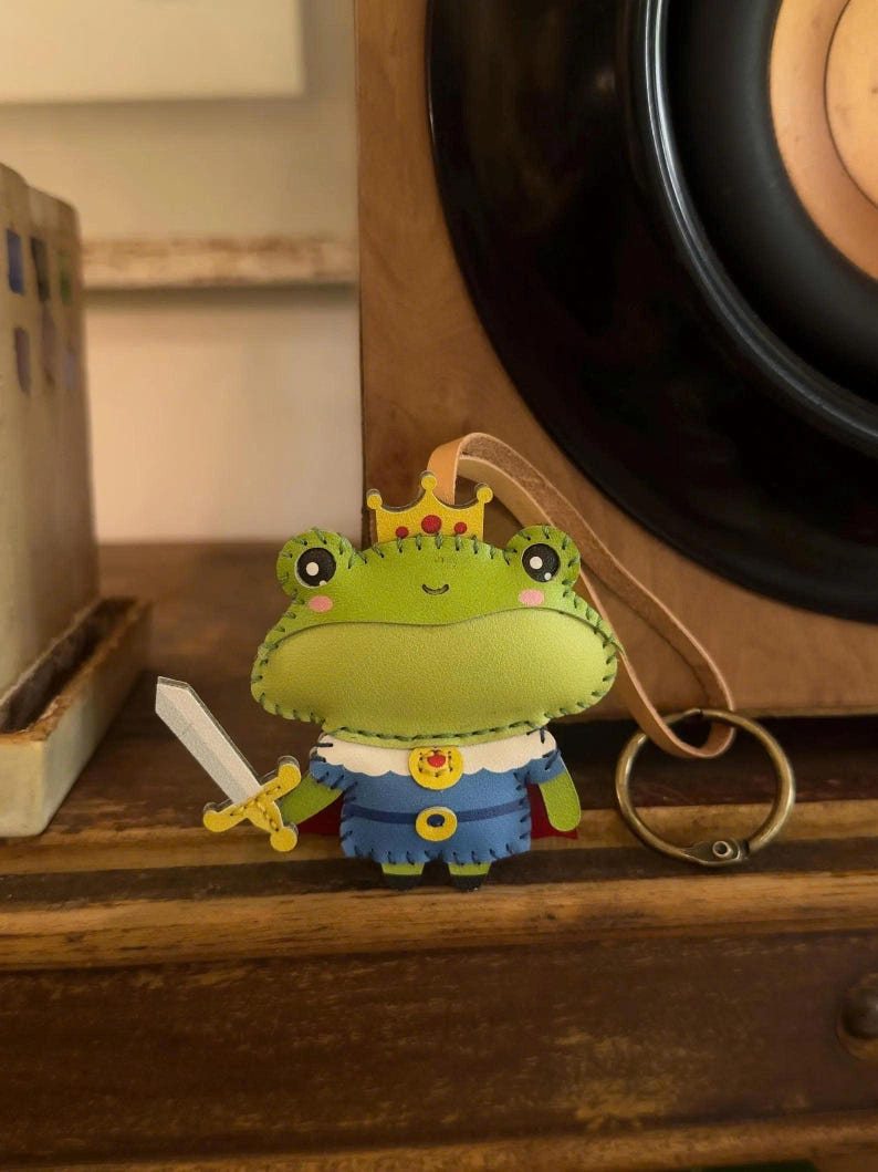May include: A handmade felt frog ornament, wearing a blue and gold outfit with a red cape and a gold crown. The frog holds a silver sword. The ornament has a leather loop for hanging and is set against a wooden background.